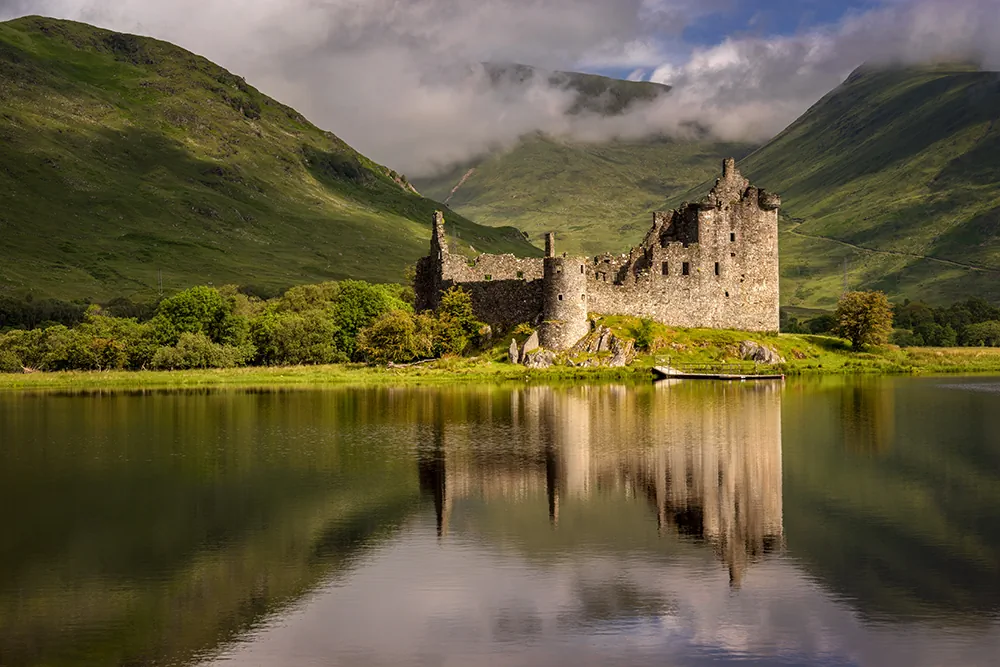 A historic castle sits beside a tranquil lake, with rugged mountains rising in the distance